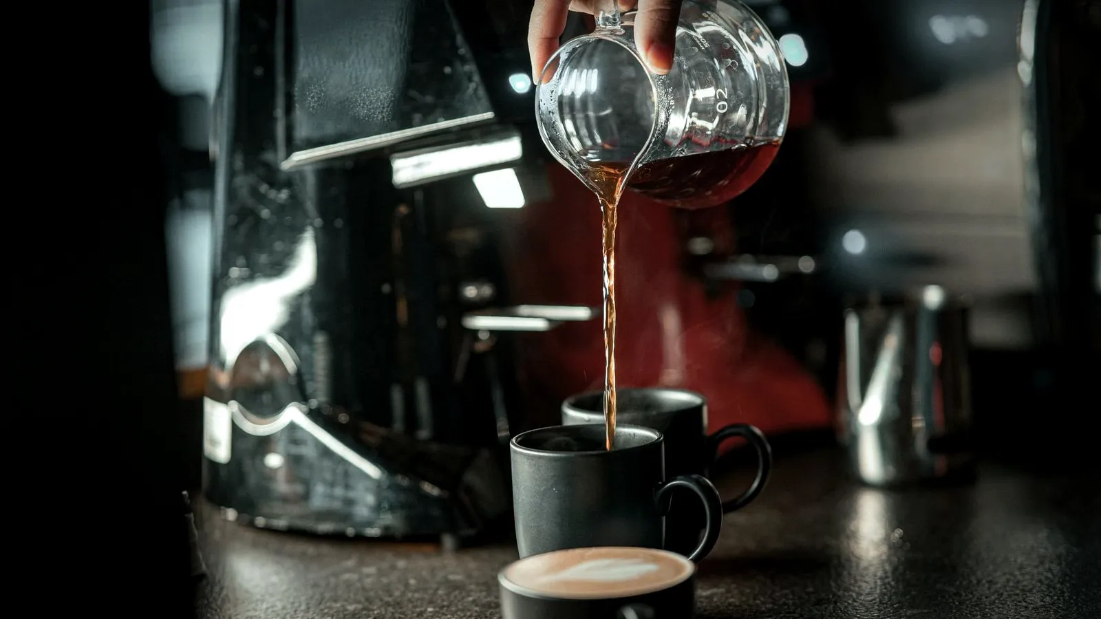 Brewed coffee poured from a glass server into a dark mug on a café counter, with a latte in front