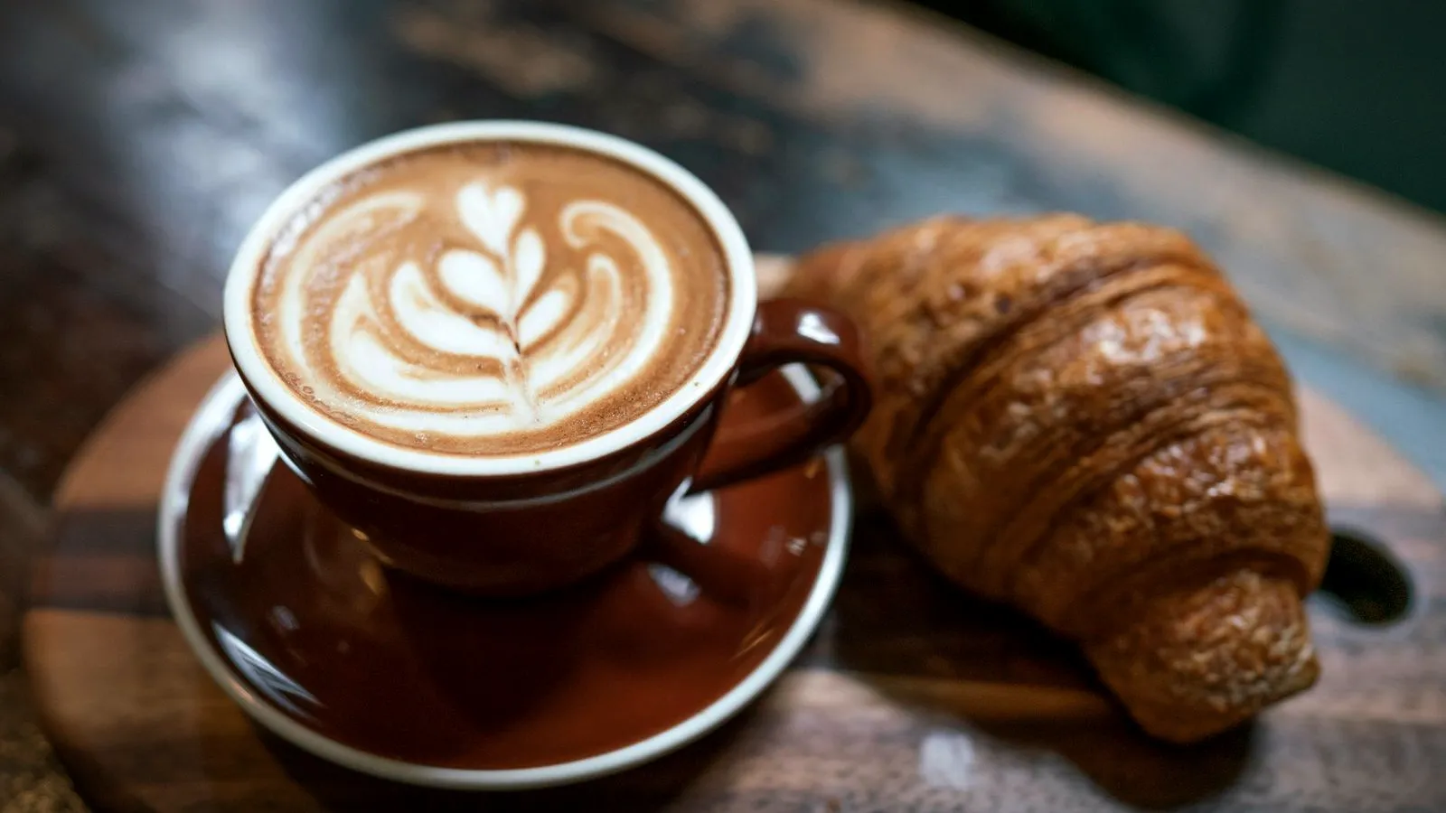 A cappuccino with latte art in a brown cup beside a croissant on a wooden board