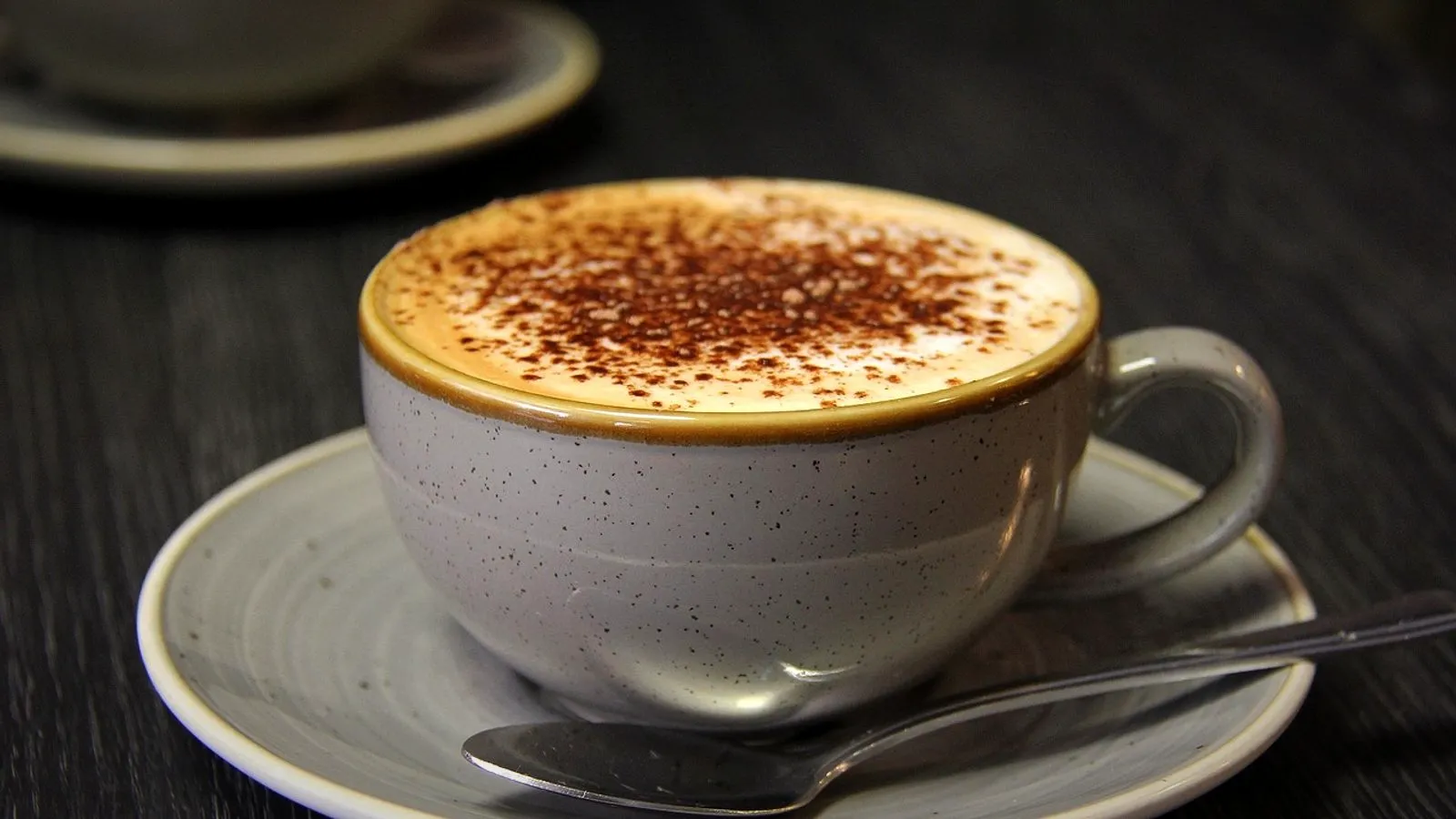 Cappuccino with cocoa-dusted foam in a grey ceramic cup on a saucer with a spoon