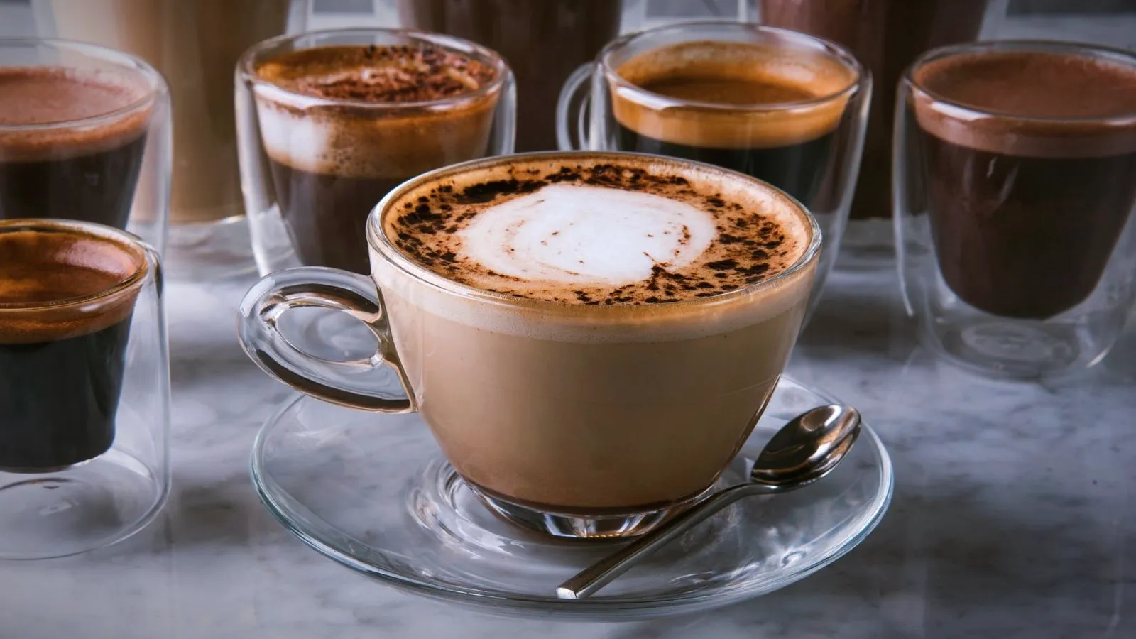 A cappuccino dusted with cocoa in a glass cup, surrounded by small glasses of espresso on a marble counter