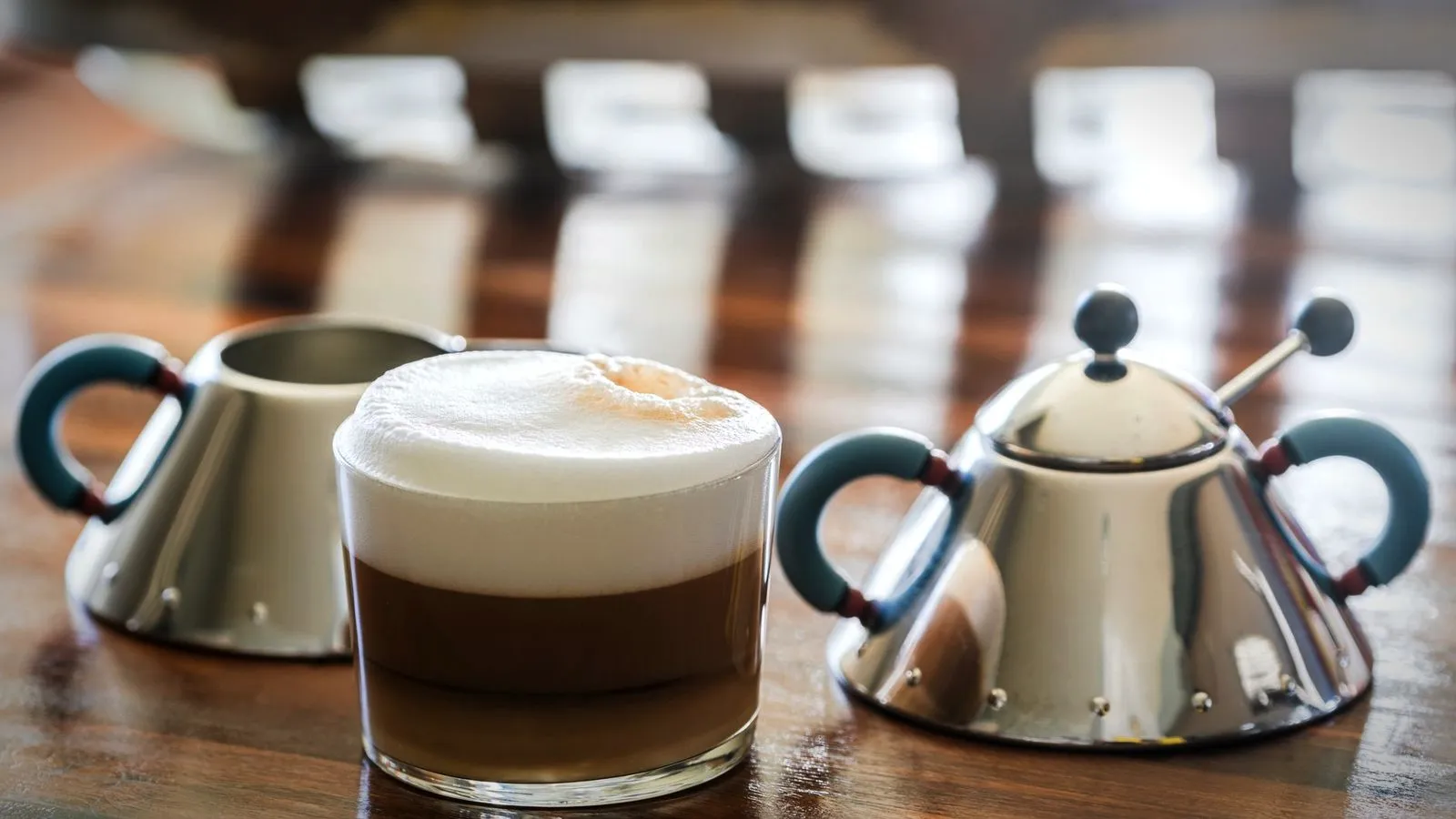 Cappuccino in a clear glass with a tall dome of milk foam over the coffee, next to a steel creamer and sugar bowl