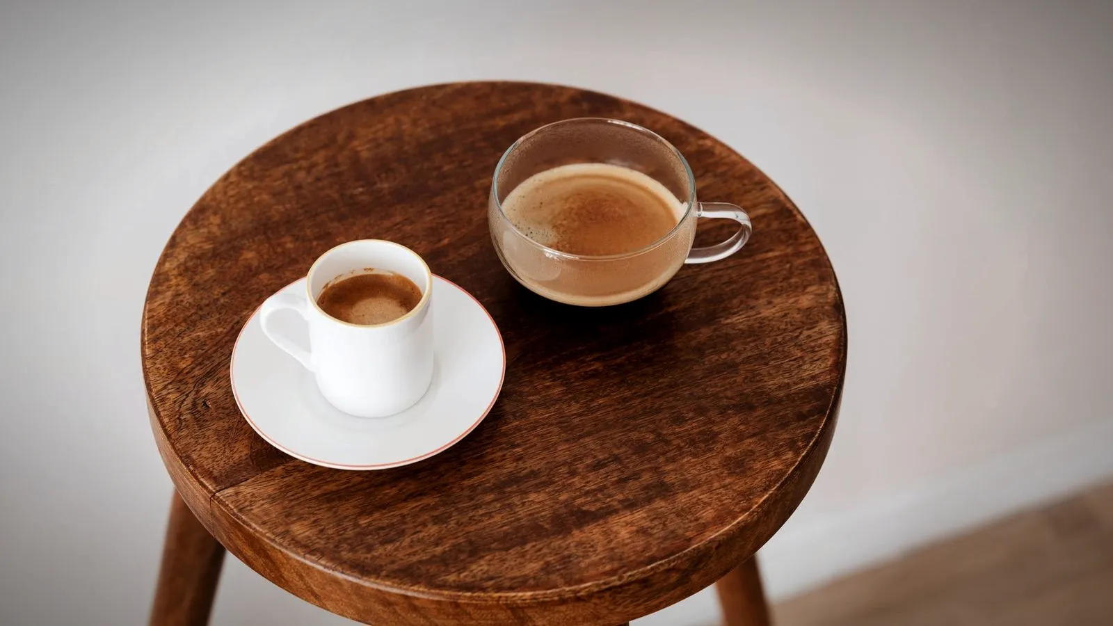 A white ceramic espresso cup beside a small clear glass cup of coffee on a wooden stool
