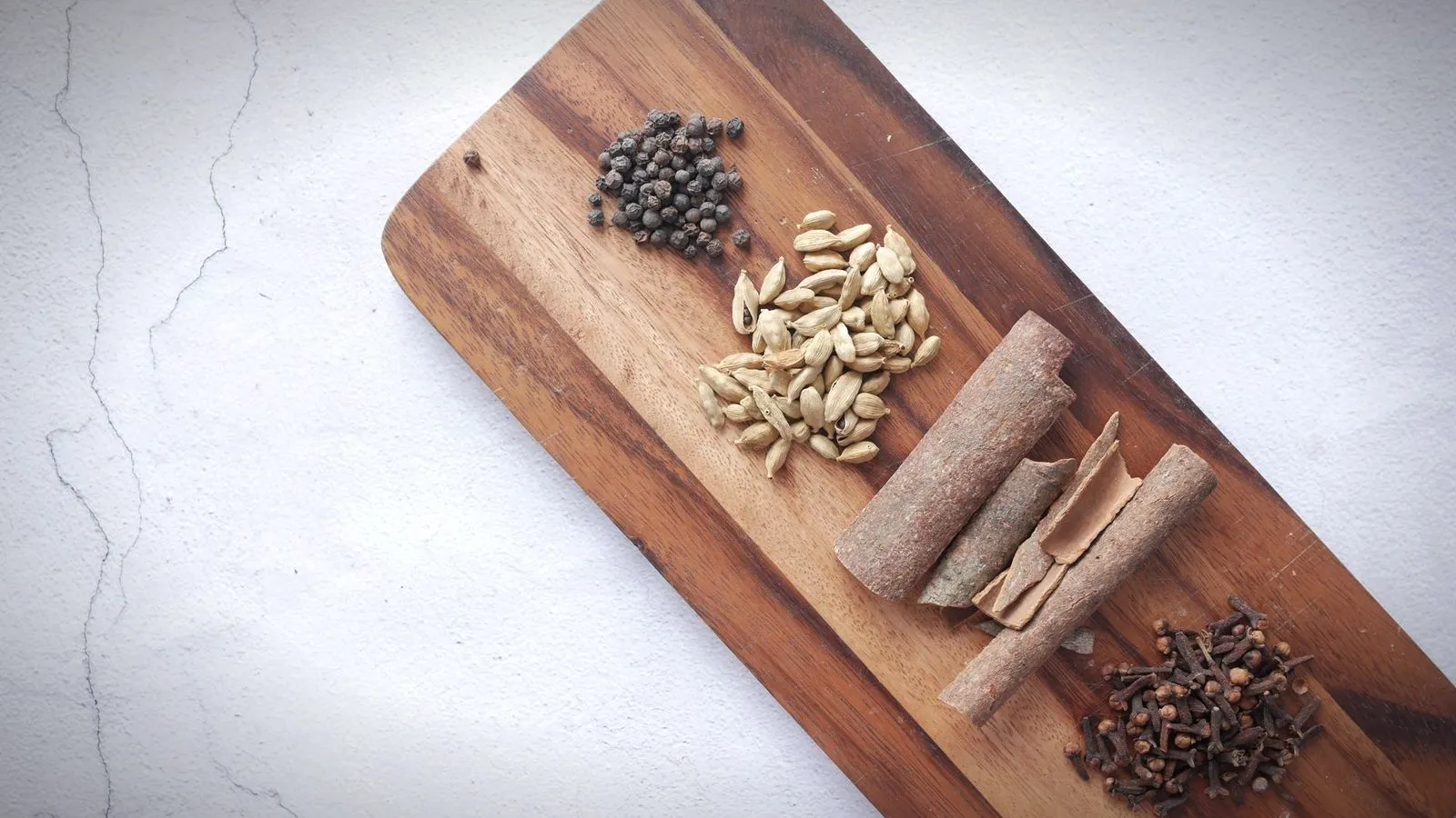 Black peppercorns, green cardamom pods, cinnamon sticks and cloves arranged on a wooden board
