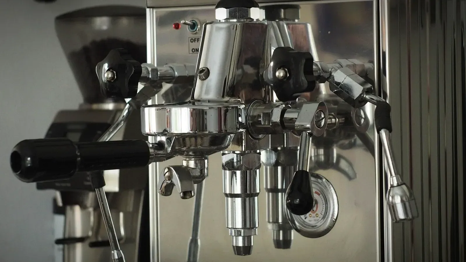 Chrome espresso machine with an E61-style group head, a portafilter locked in and a small pressure gauge, with a grinder behind it