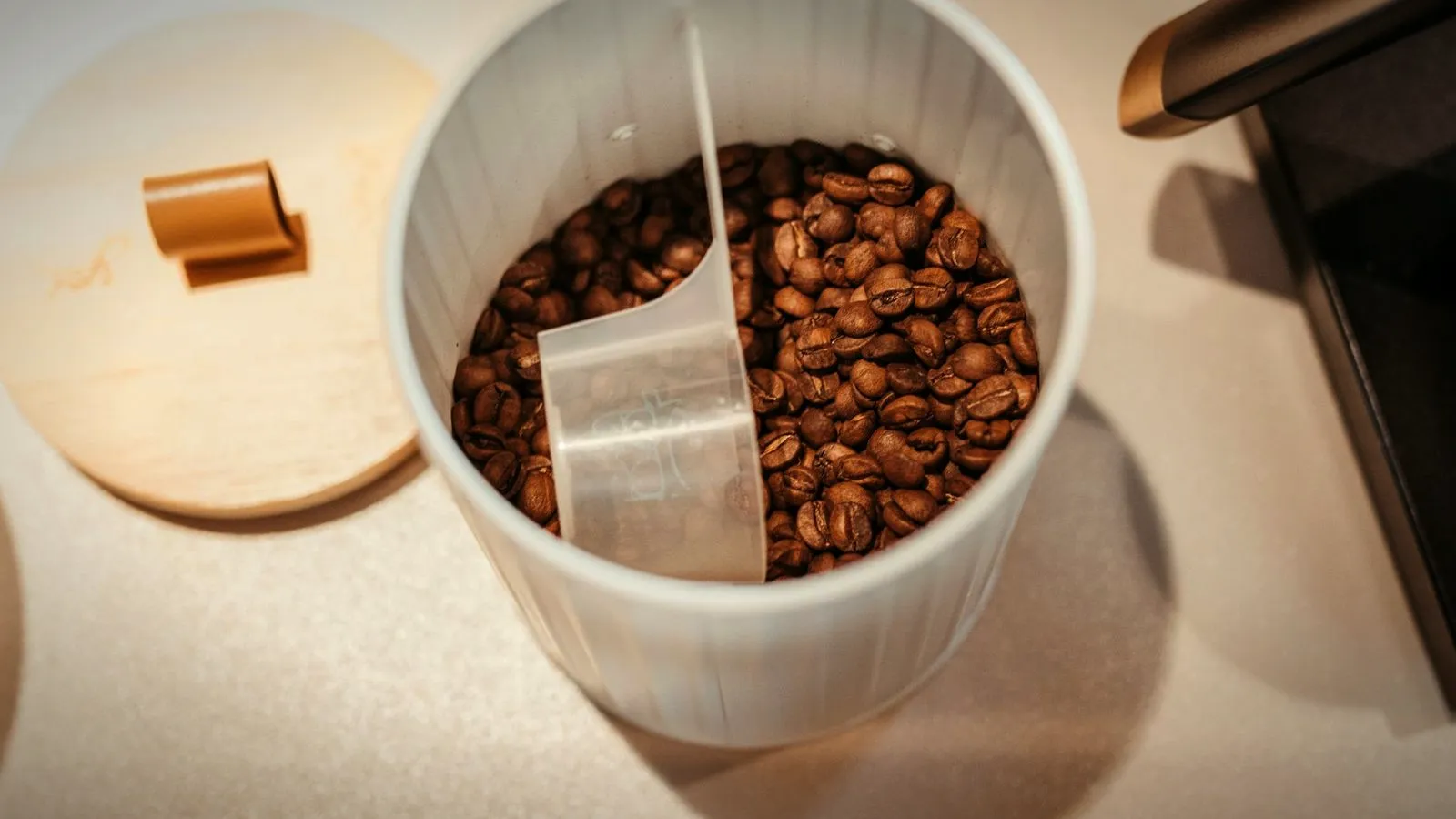Roasted coffee beans in a white canister with a measuring scoop, its wooden lid set aside