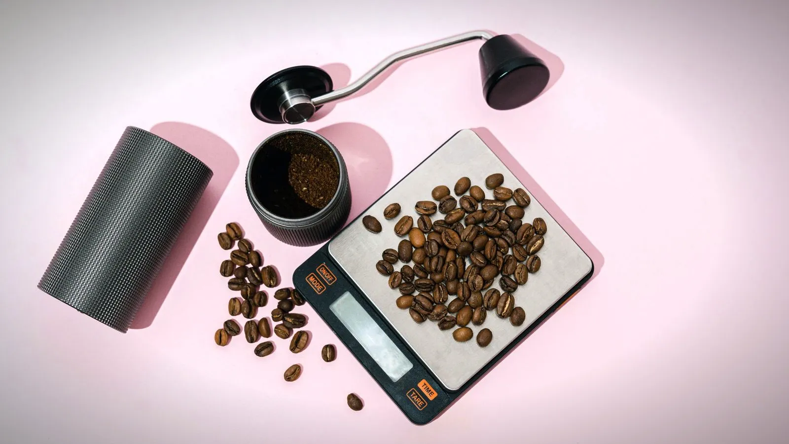 Top view of roasted coffee beans on a digital scale with time and tare buttons, next to an opened hand grinder