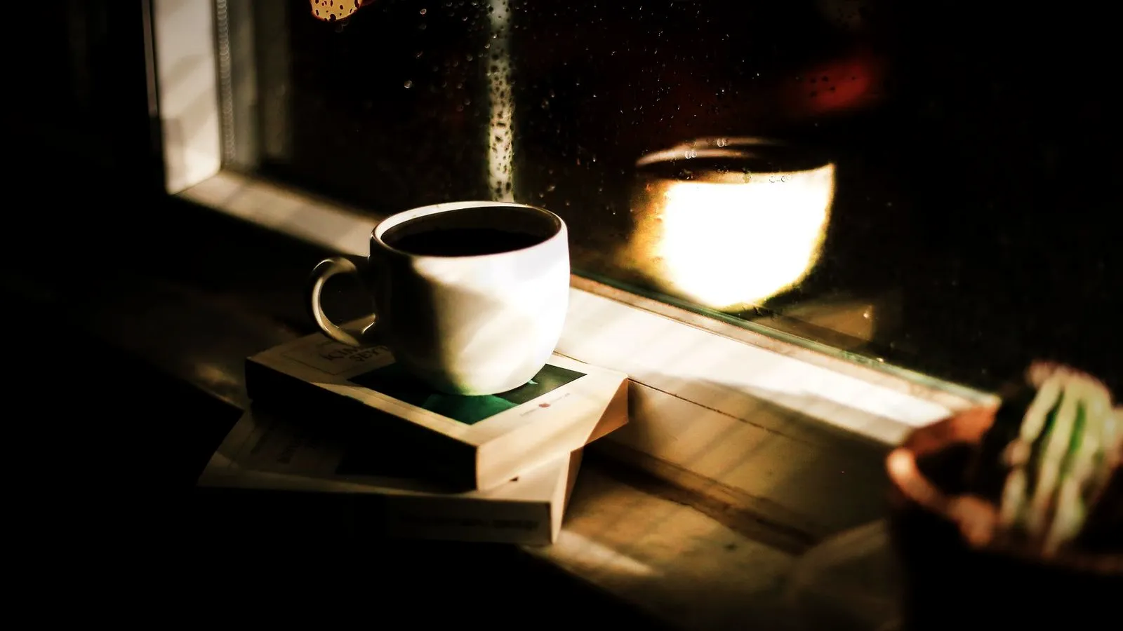 A cup of coffee on a stack of books by a rainy window at night