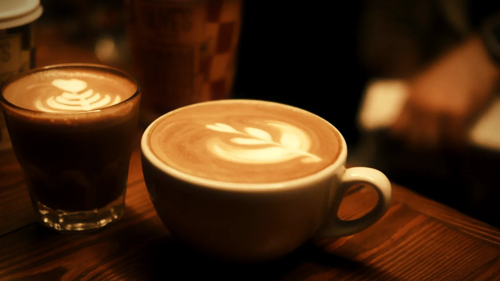A cortado in a small glass beside a latte with latte art in a large cup on a wooden table