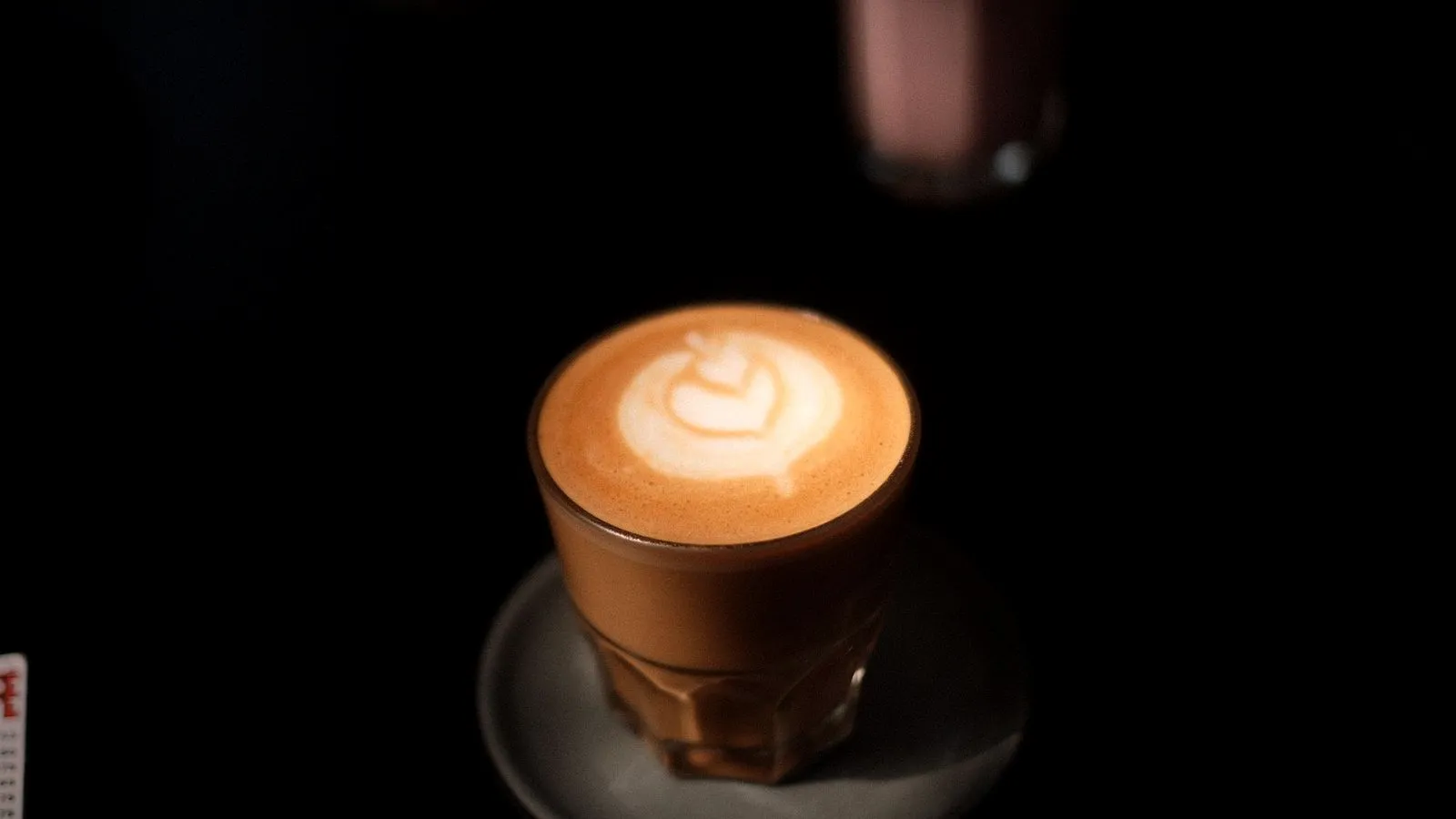 A cortado with a small latte art rosetta in a short glass on a saucer against a dark background