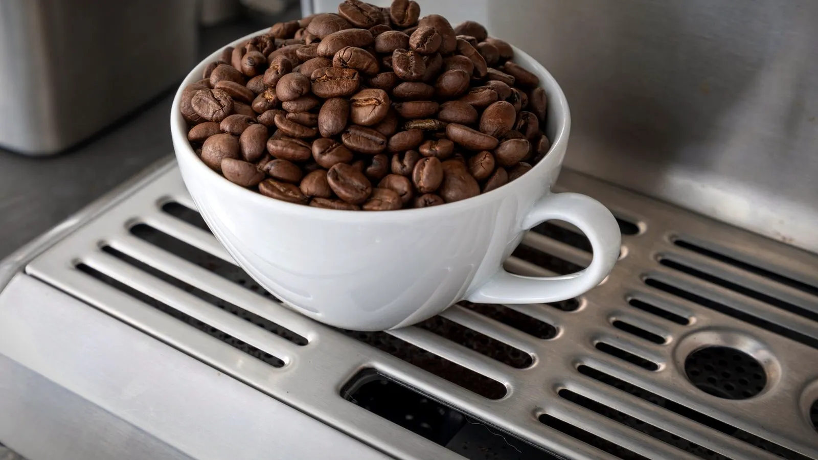 A white cup full of roasted coffee beans on the drip tray of a coffee machine