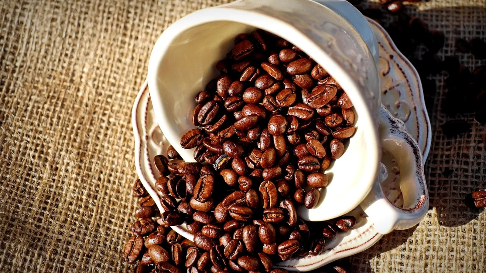 A tipped white cup spilling roasted coffee beans onto burlap