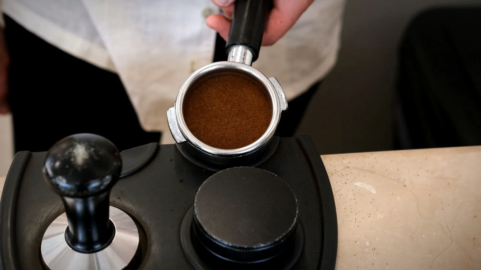Portafilter filled with an even bed of ground coffee held above a tamping station next to a tamper