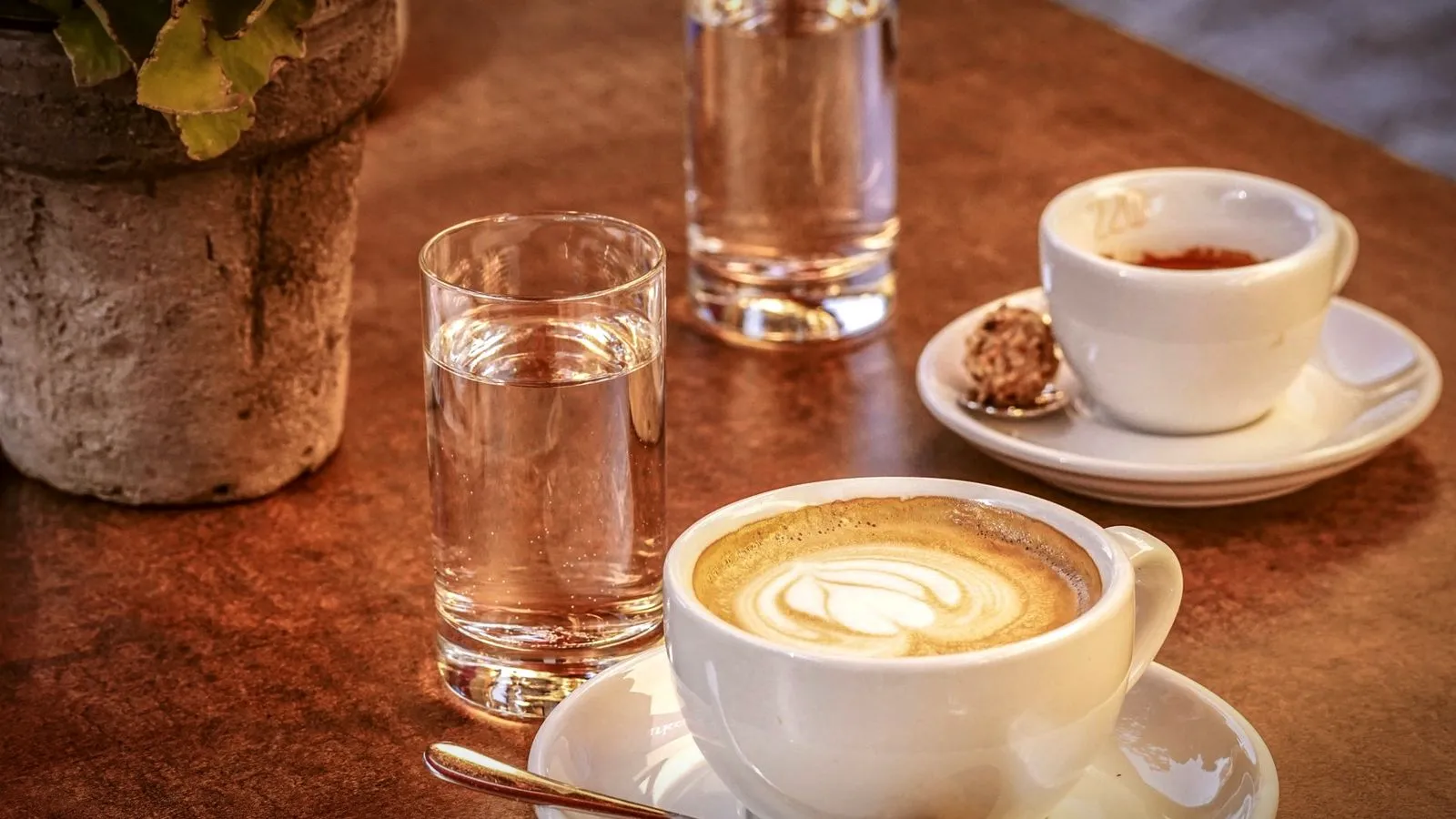 A small espresso cup next to a larger cappuccino cup and glasses of water on a café table