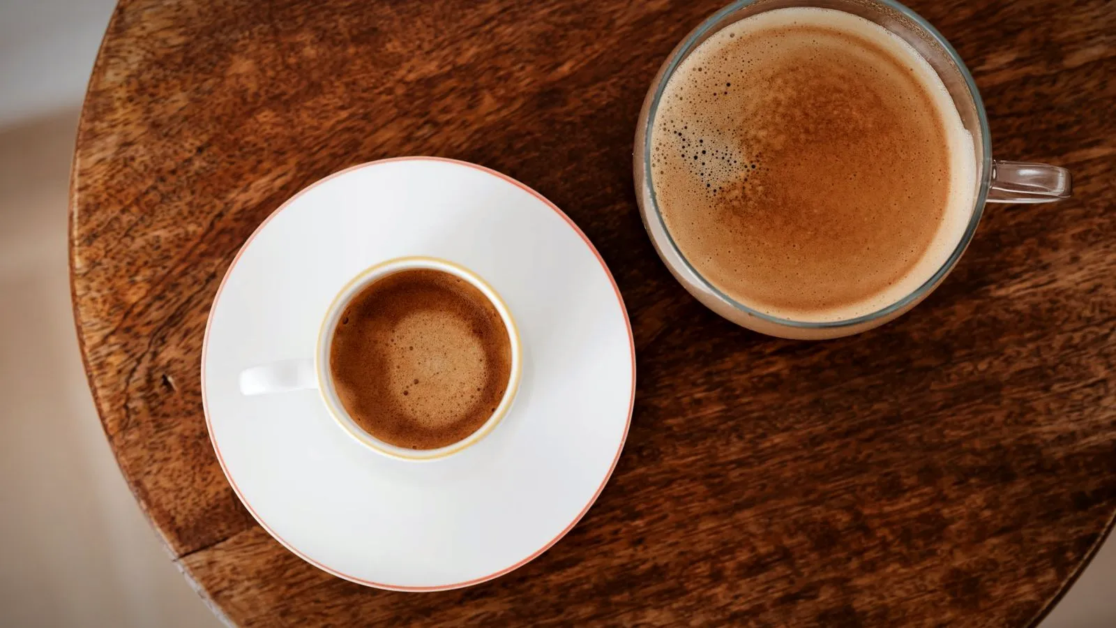 Top view of a small cup of espresso on a saucer beside a glass of milky coffee on a round wooden table