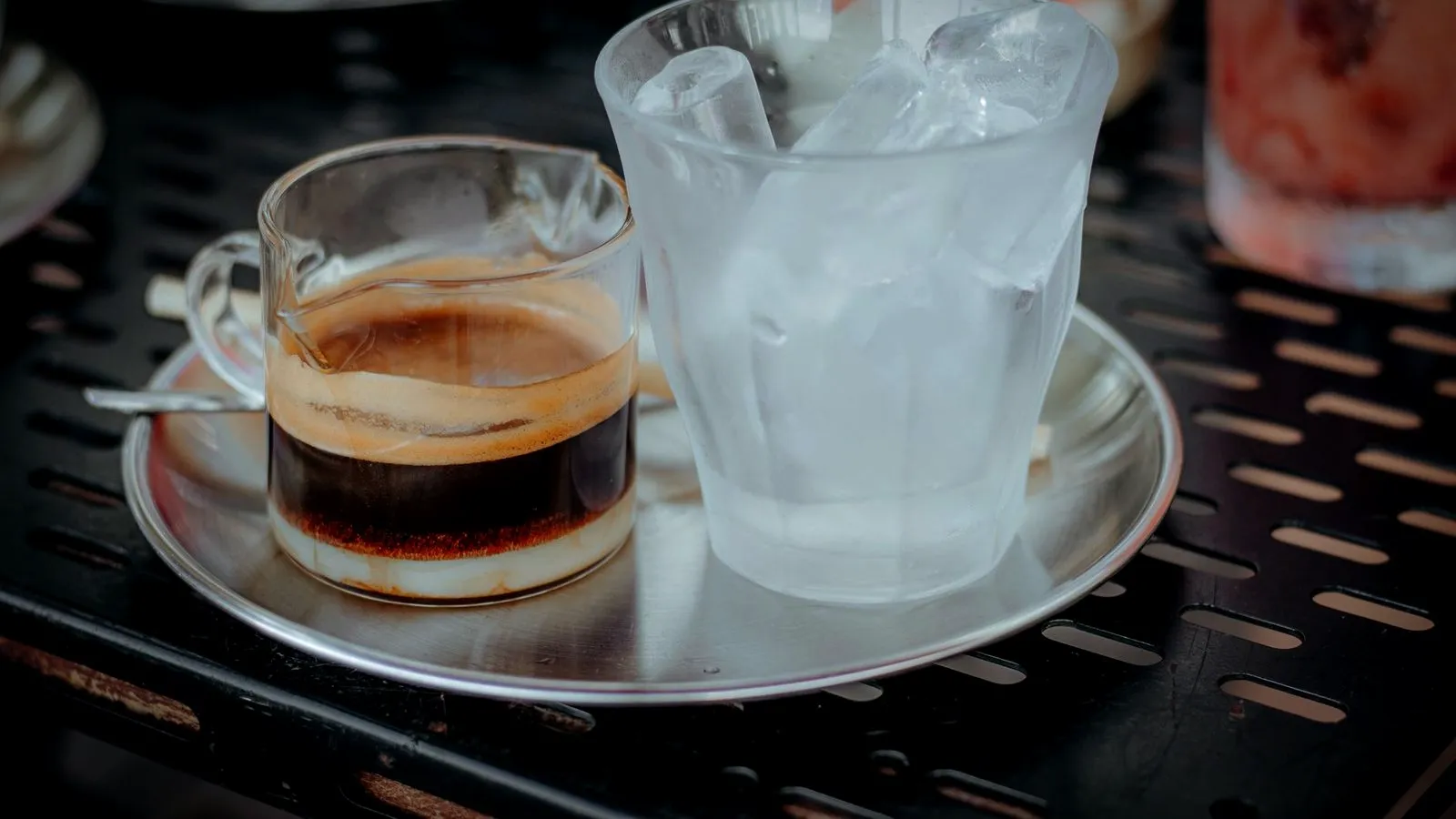 A glass cup of espresso beside a tall glass of ice water on a metal café table