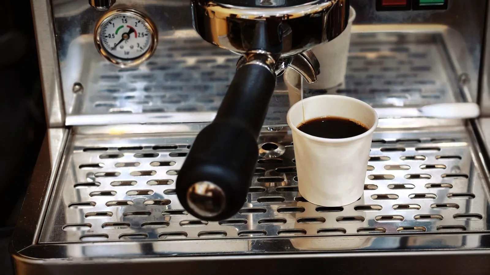 Small 0 to 3 bar boiler pressure gauge on an espresso machine, next to a locked-in portafilter and a paper cup of coffee on the drip tray