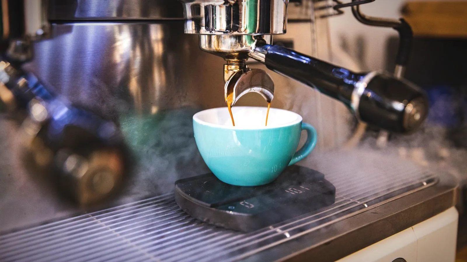 Espresso pouring from a portafilter into a turquoise cup on a small scale, with steam around the group head