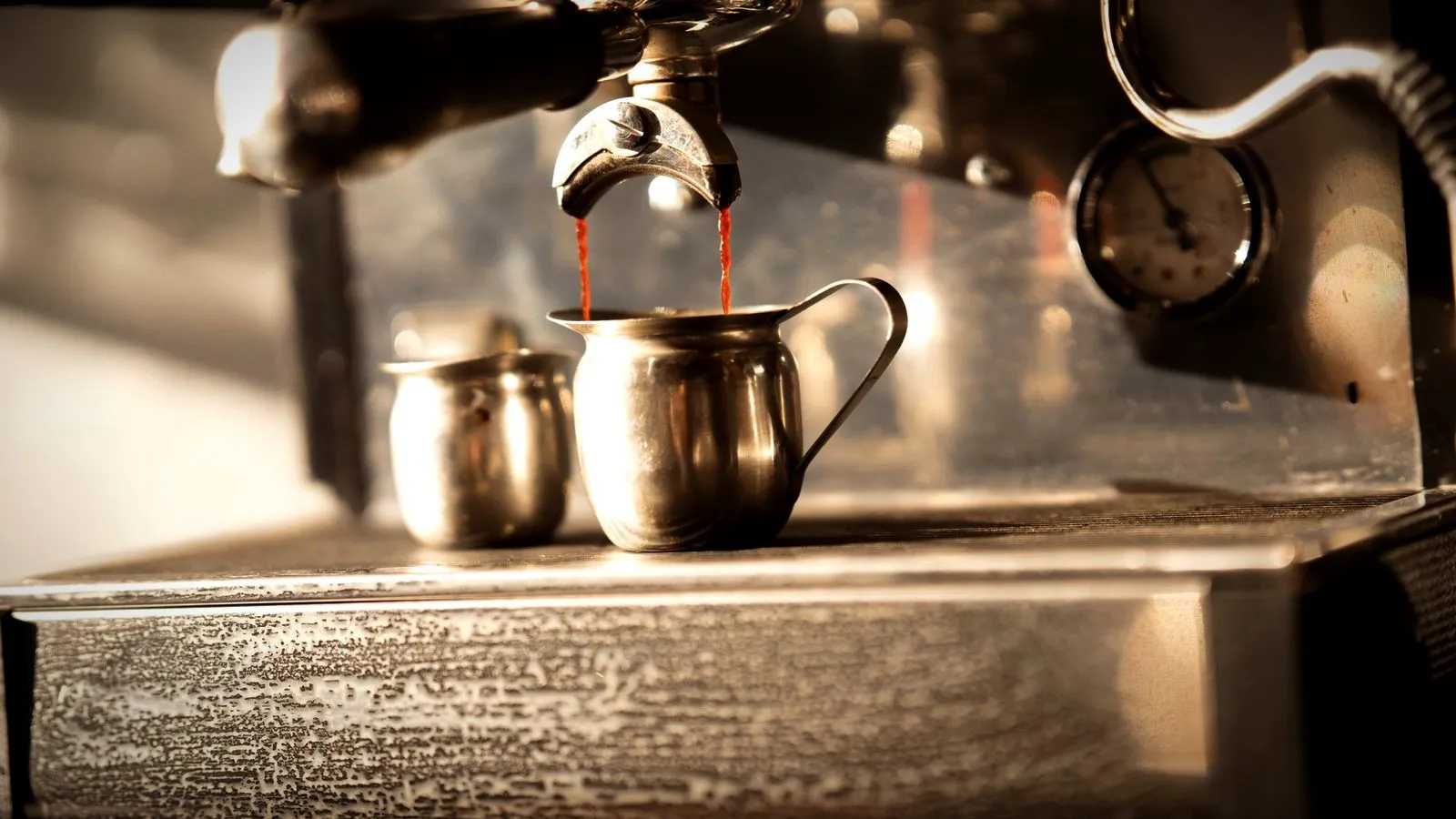Espresso pouring from a spouted portafilter into a small steel pitcher, with a second pitcher beside it and a round pressure gauge on the machine