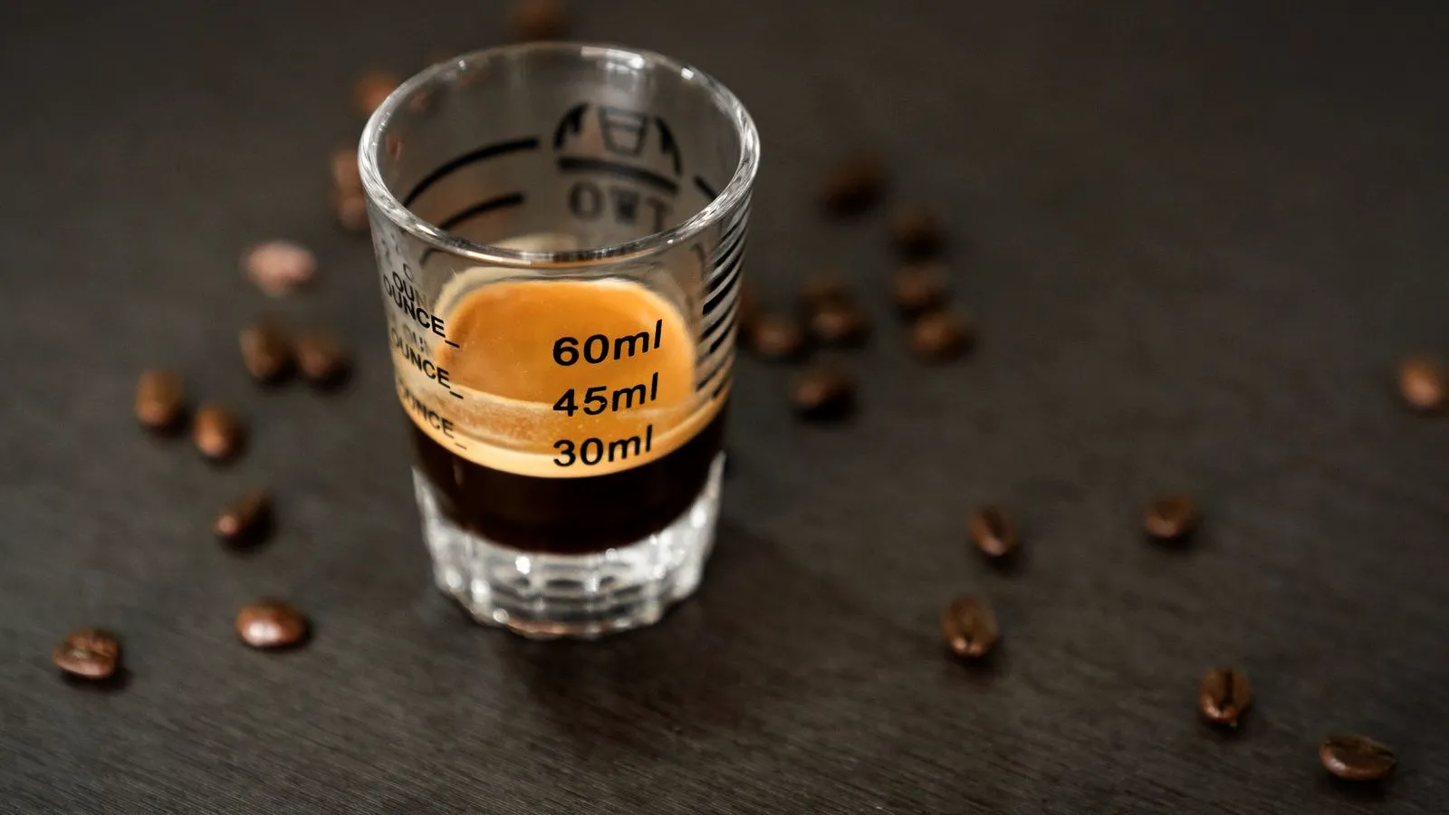 Single espresso shot in a measuring shot glass marked 30, 45 and 60 ml, with roasted coffee beans scattered around