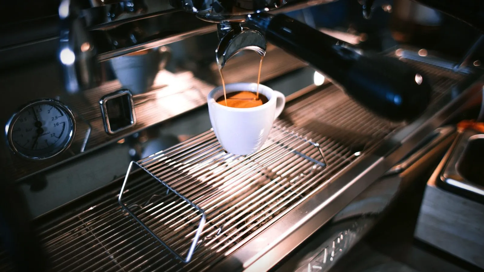 Espresso flowing into a white cup on a stainless drip tray, with the machine’s pressure gauge beside it
