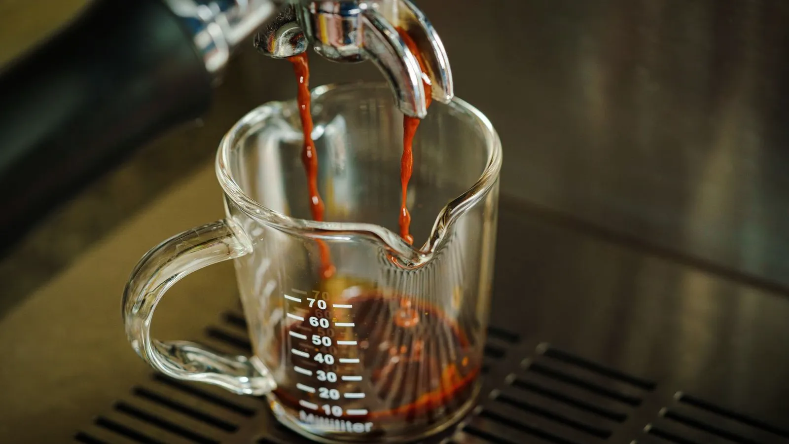 Two streams of espresso running into a glass shot pitcher with measurement marks on the drip tray