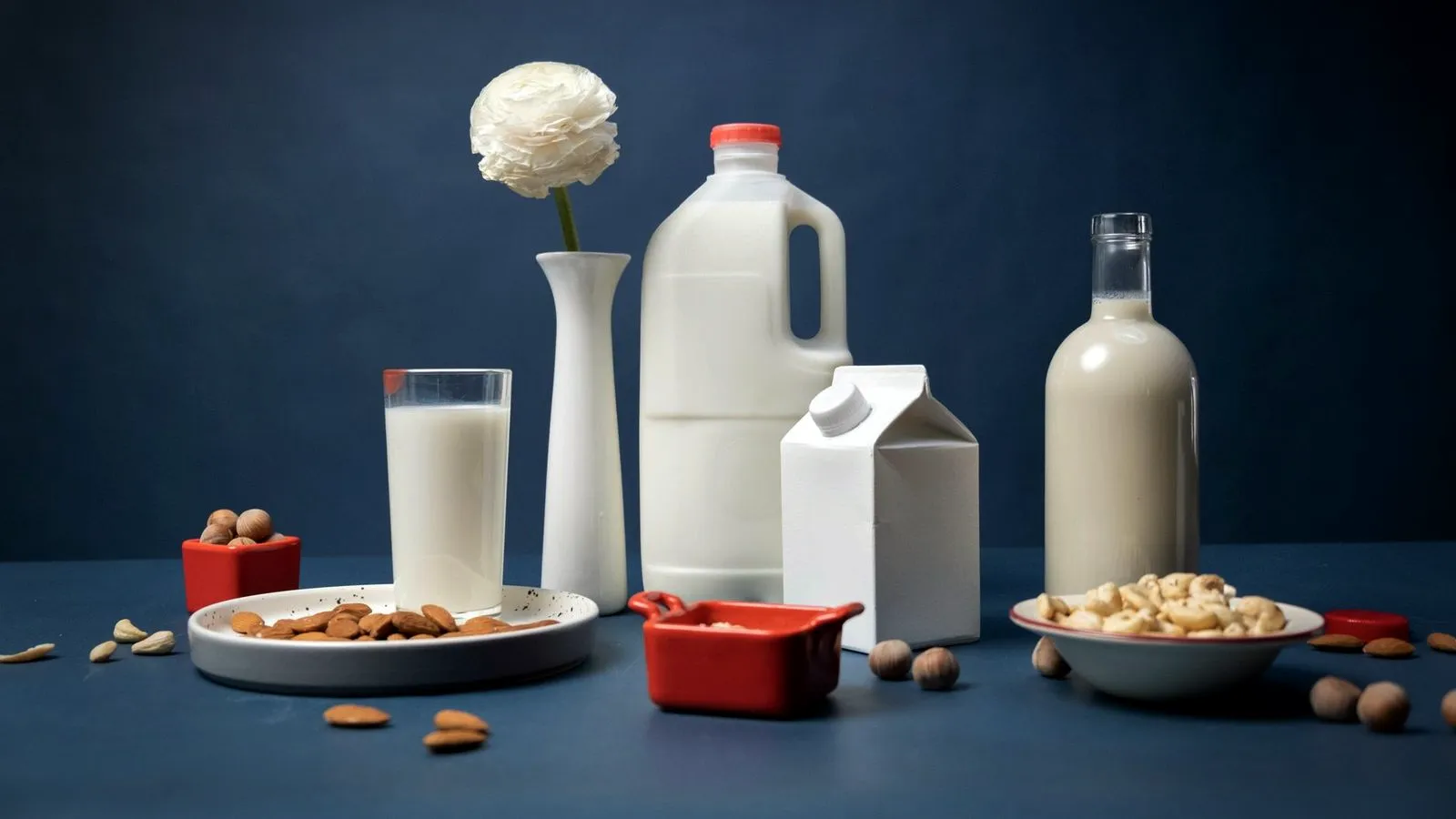 Plain white milk jug, carton and glass bottle with a glass of milk and bowls of almonds, hazelnuts and cashews on a blue table