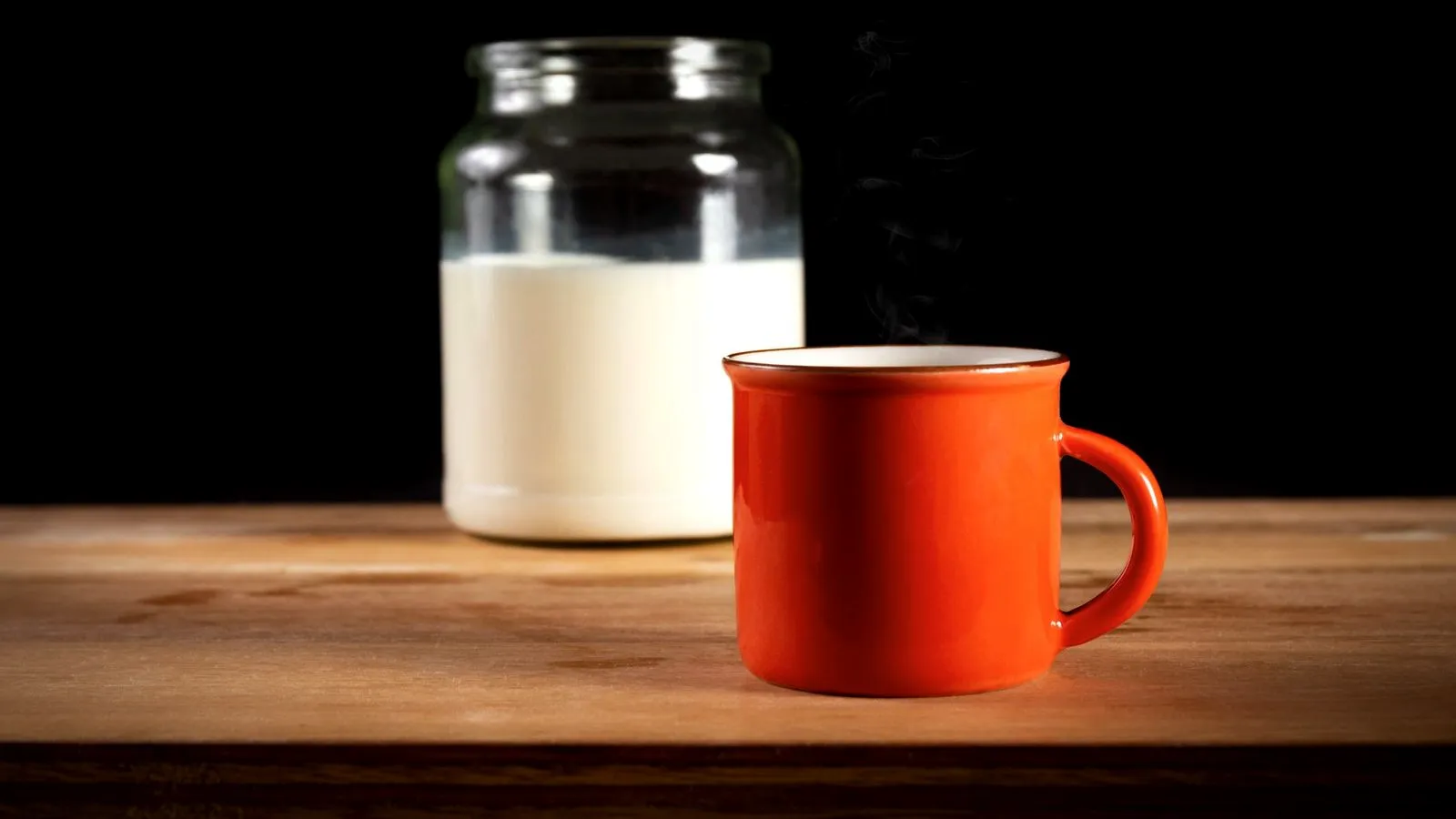 A glass jar of milk next to a steaming red enamel mug on a wooden table