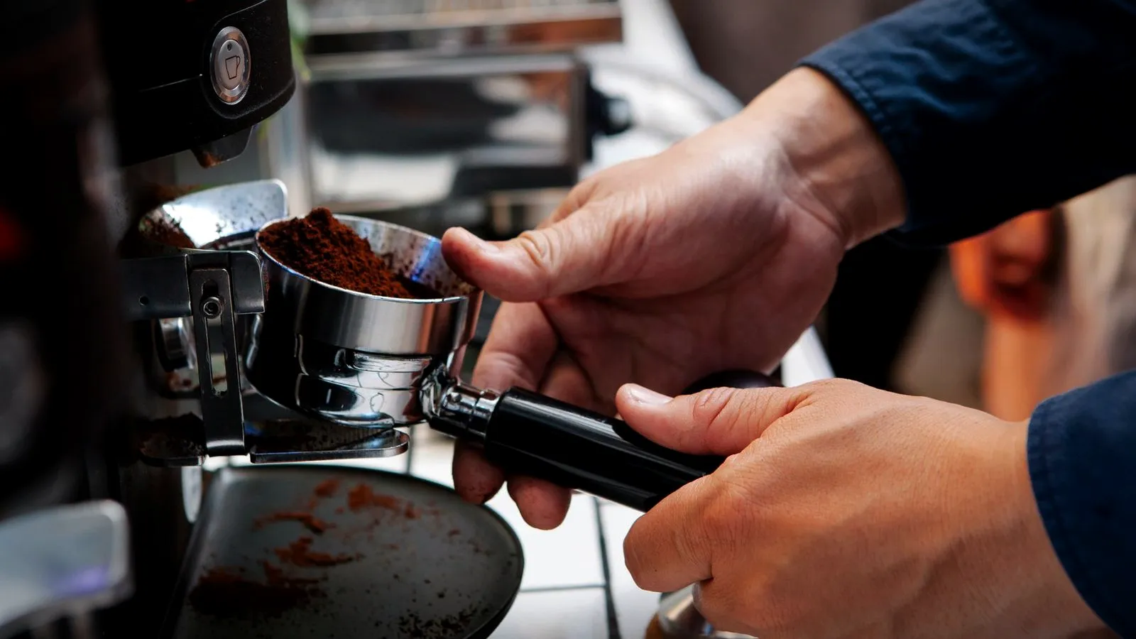 Hands holding a portafilter under a grinder spout as it fills with a heap of fresh grounds
