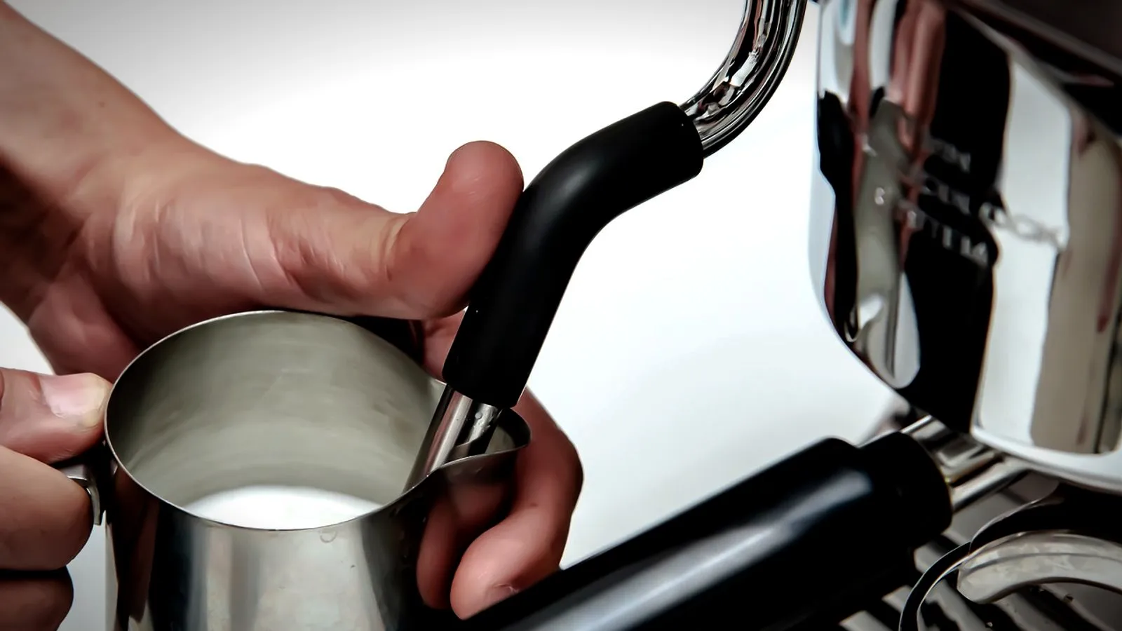 Hand holding a steel milk pitcher under an espresso machine steam wand