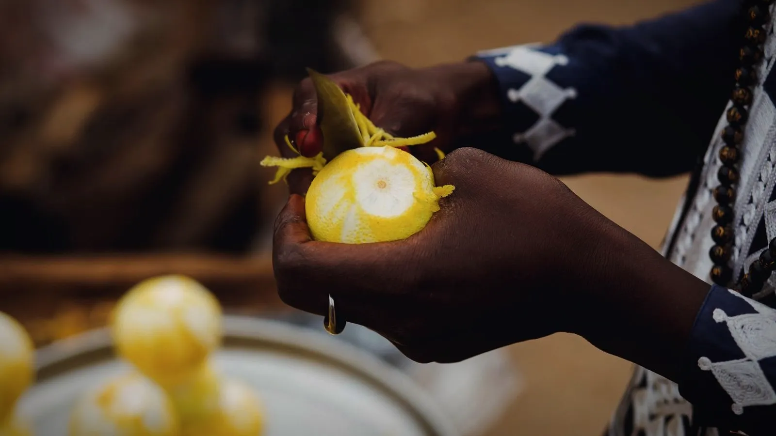 Hands peeling thin strips of yellow zest from a lemon with a small knife, with peeled lemons on a plate