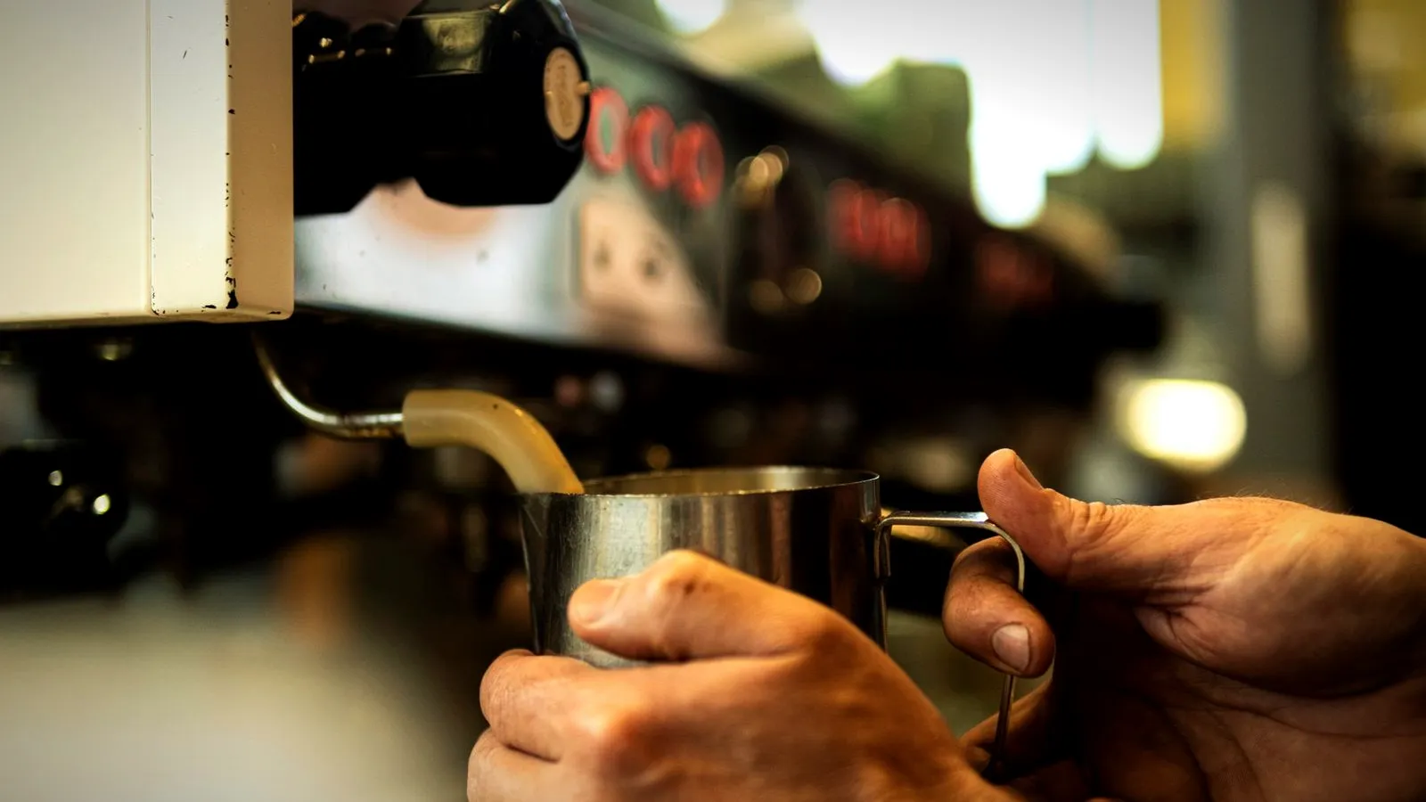 Hands holding a stainless steel pitcher under an espresso machine’s steam wand while texturing milk