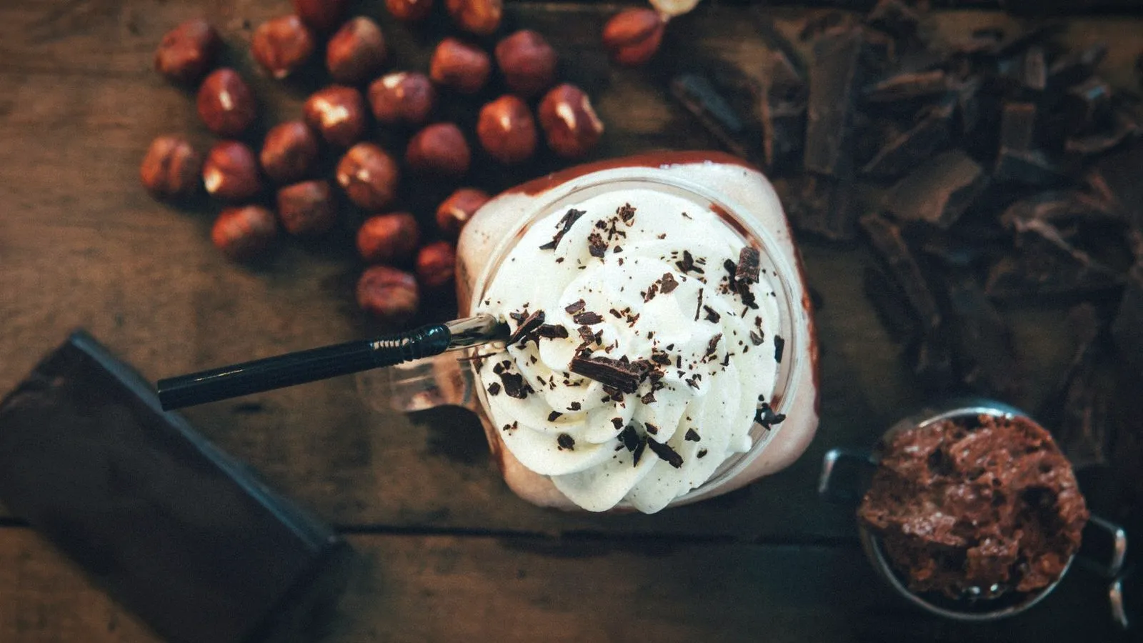 A coffee drink in a jar glass topped with whipped cream and chocolate shavings, surrounded by whole hazelnuts and chocolate pieces
