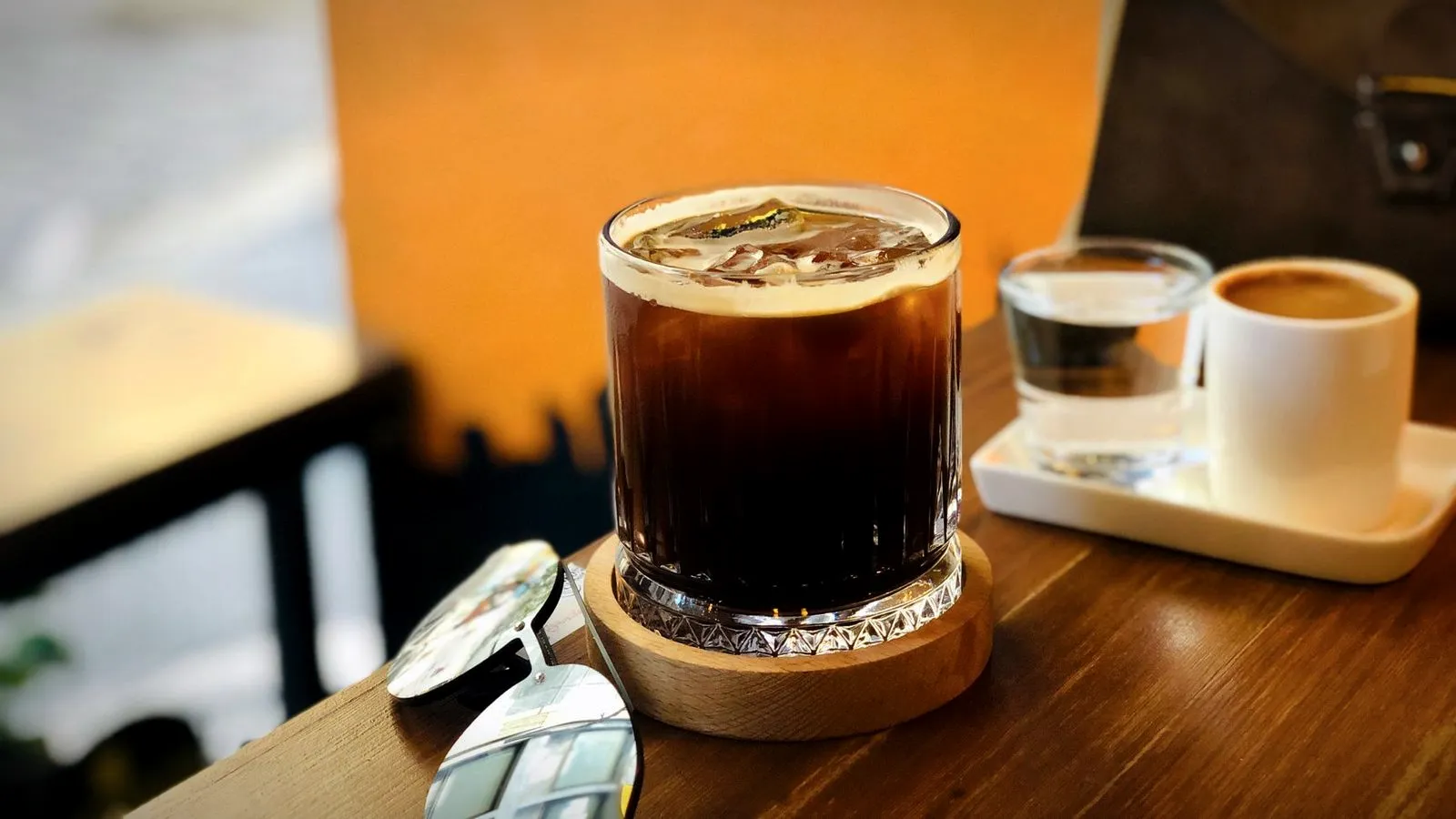 An iced Americano in a short glass on a wooden coaster beside a small glass of water and a cup of espresso on a tray