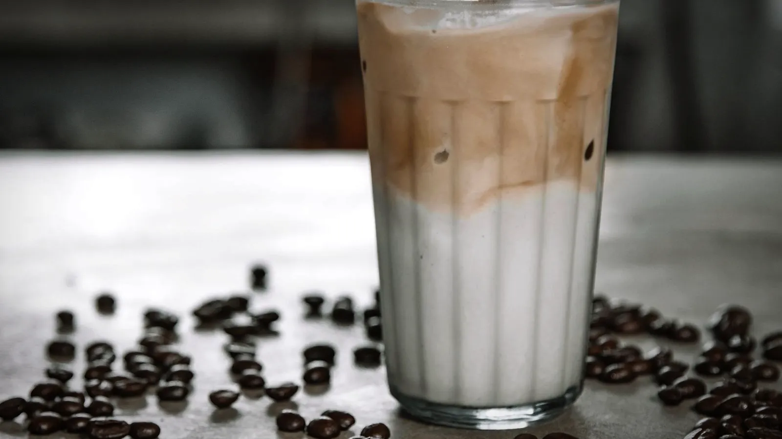 An iced latte in a tall glass with espresso swirling into the milk, coffee beans scattered on the table
