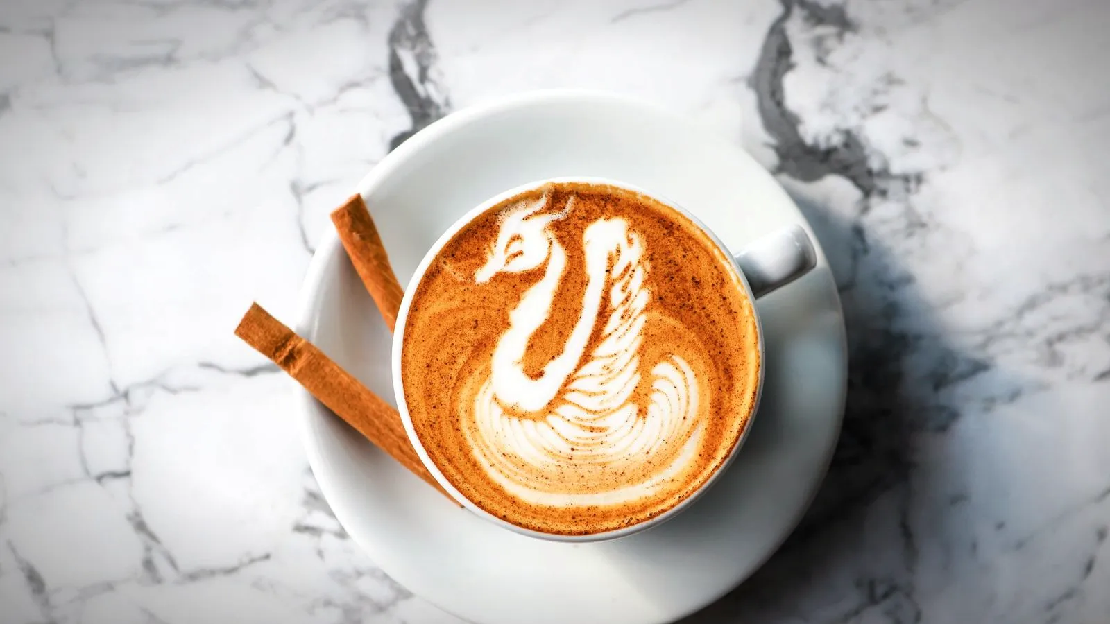 Latte with swan latte art on a white saucer with two cinnamon sticks, on a marble surface