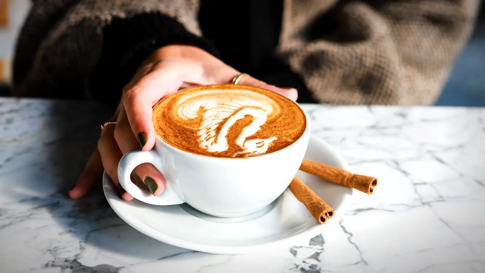 Hand holding a latte with swan latte art, two cinnamon sticks on the saucer, on a marble table