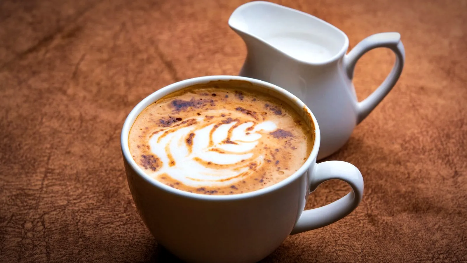 A latte with leaf-pattern latte art in a white cup beside a small jug of milk