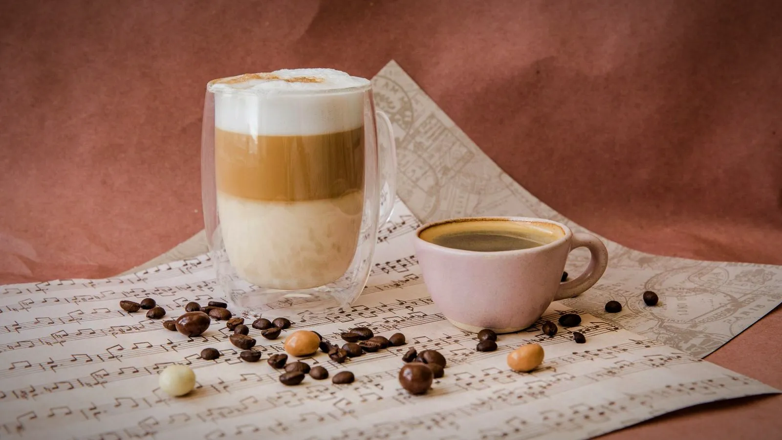 A tall layered latte in a double-walled glass beside a small cup of espresso, with coffee beans on the table