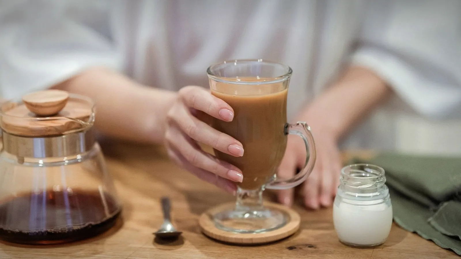Hands holding a glass mug of milky coffee on a wooden table, with a glass carafe and a small jar of milk beside it