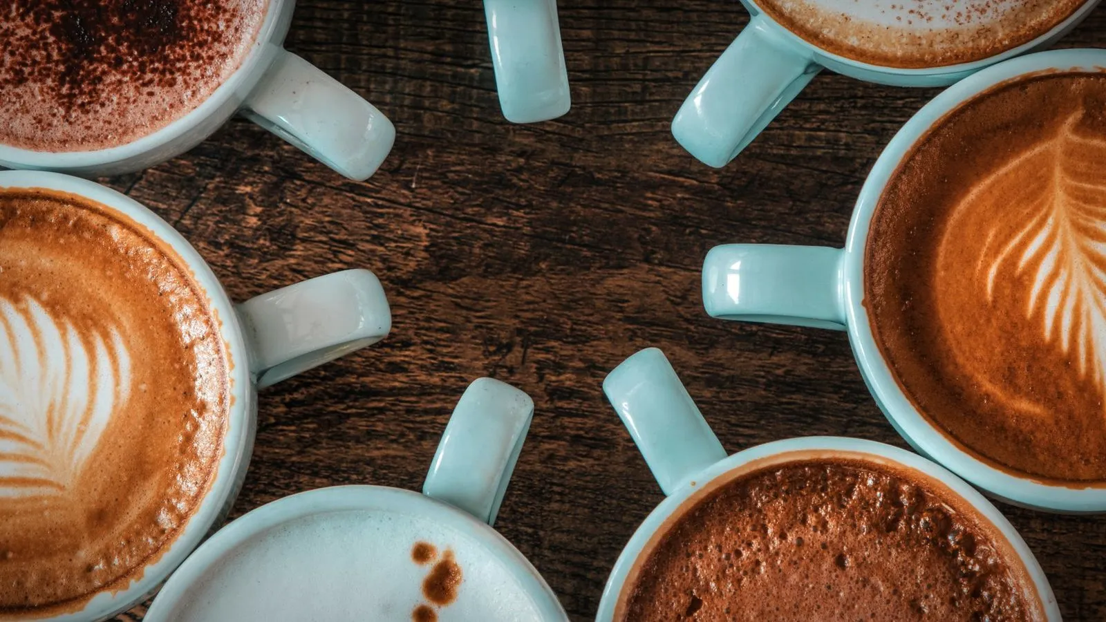 Top view of pale blue cups of milky coffee on a dark wooden table, some with latte art and one dusted with cocoa