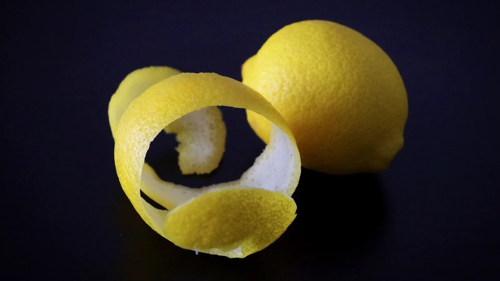 Spiral of lemon peel showing the yellow outer skin and white inner layer, beside a whole lemon on a dark background
