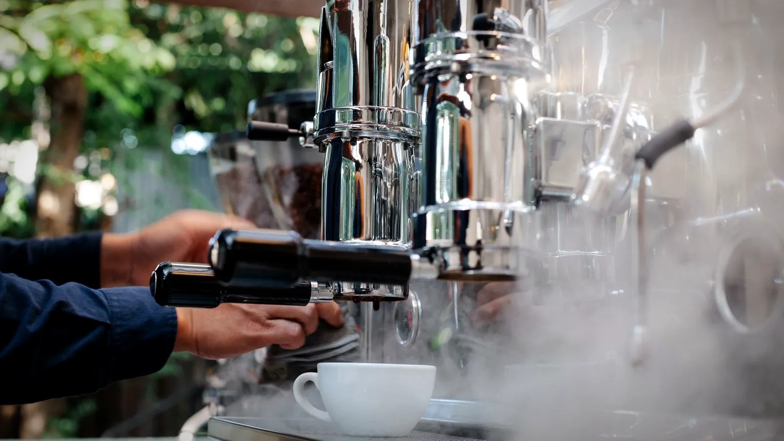 Barista at a chrome espresso machine with tall cylindrical groups, steam rising around a white cup, at an outdoor coffee stand
