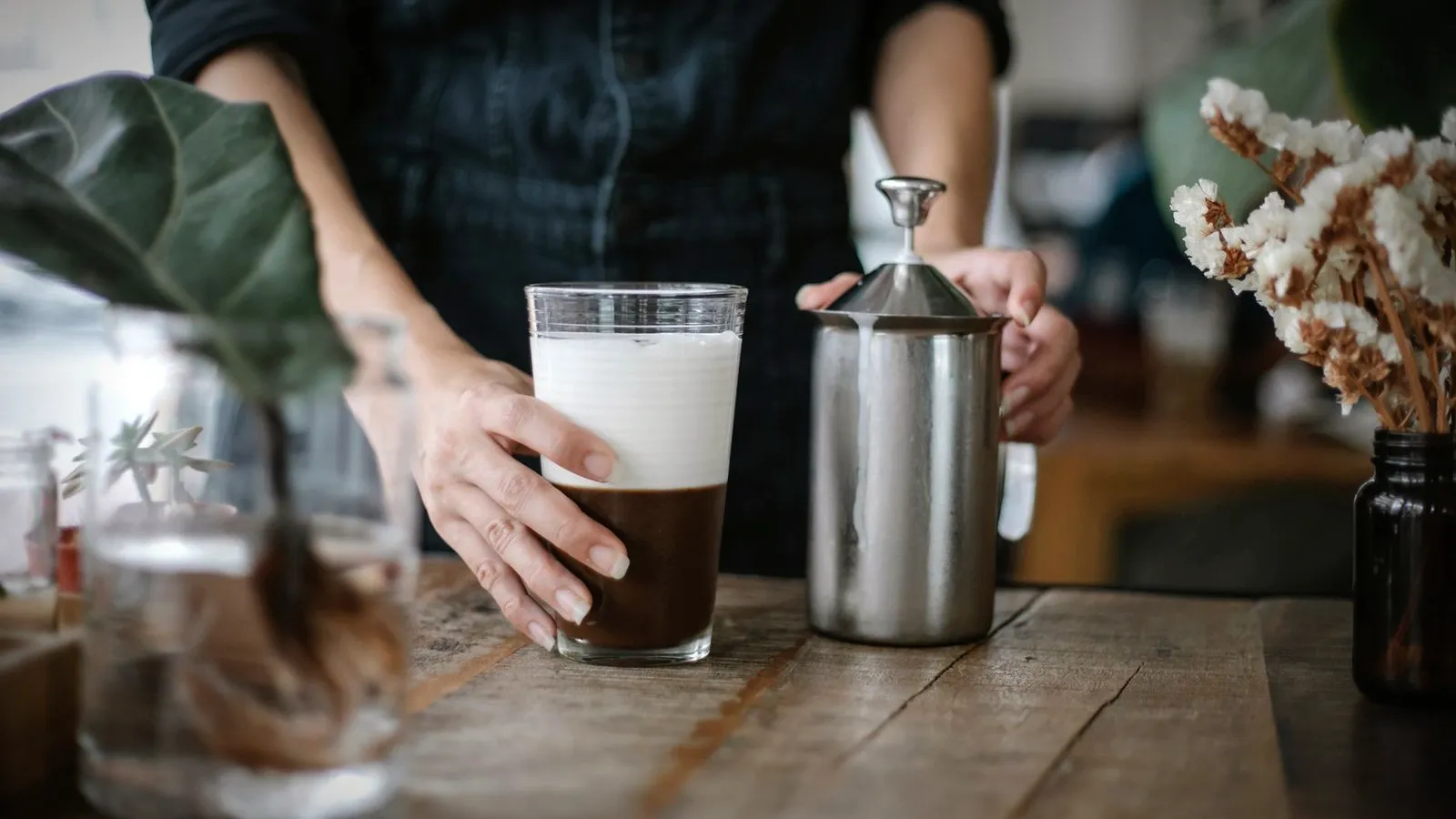 A layered coffee and milk drink in a glass next to a stainless manual milk frother