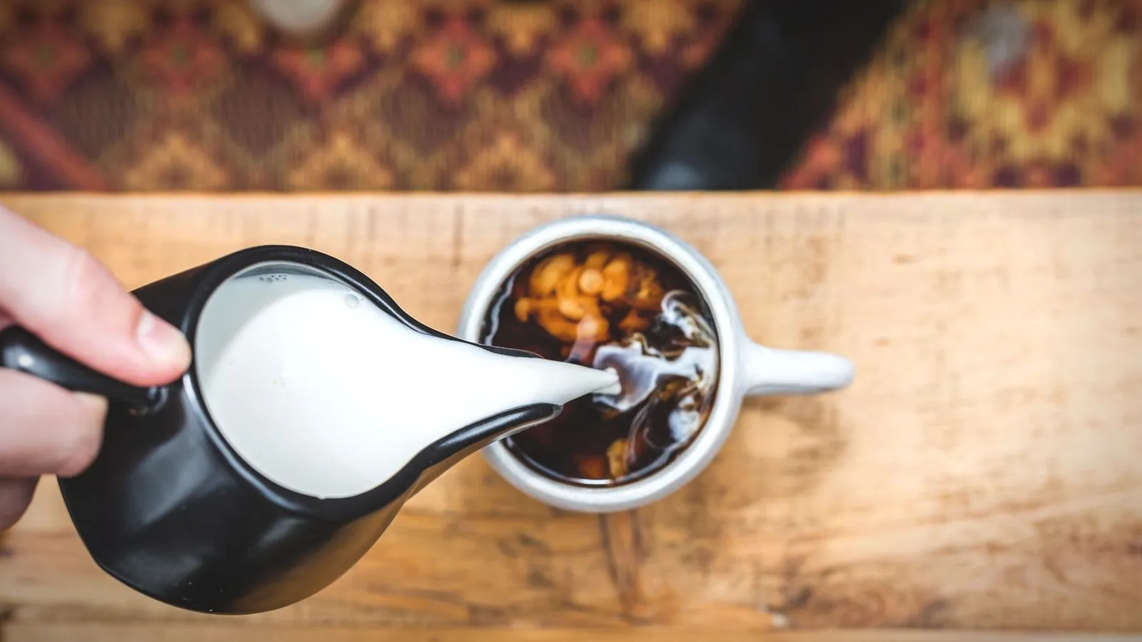 Top view of milk being poured from a small jug into a cup of black coffee on a wooden table