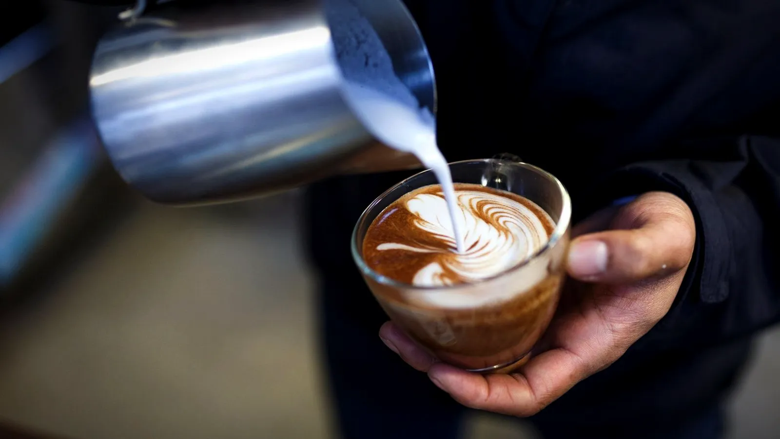 Steamed milk being poured from a pitcher into a glass cup of espresso, forming latte art