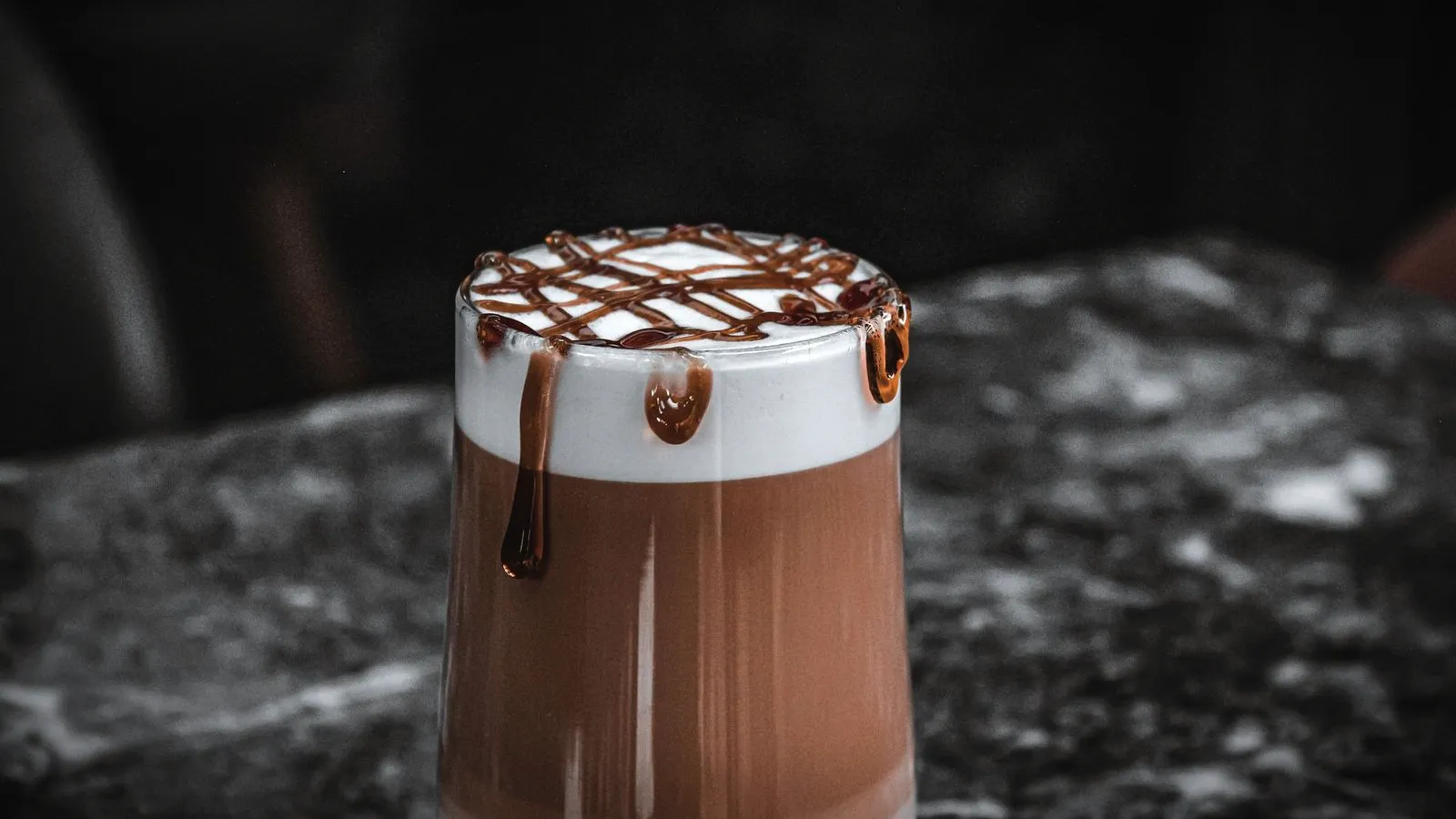 A layered mocha in a glass with chocolate sauce drizzled over the milk foam on a dark marble counter