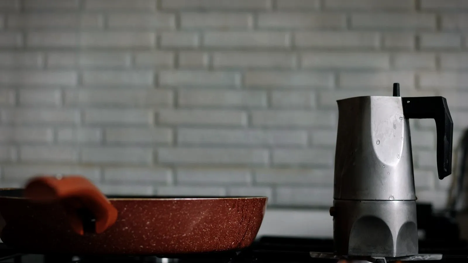 A stainless steel moka pot on a gas stove beside a frying pan