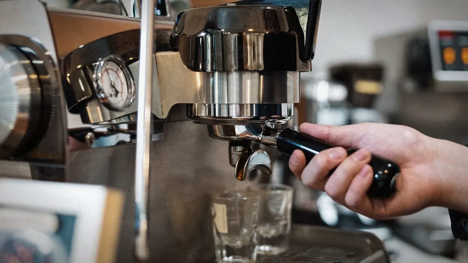 Hand on a portafilter locked into an espresso machine, with a round pressure gauge beside the group head and two shot glasses below