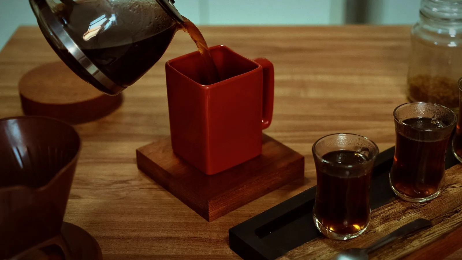 Filter coffee being poured from a glass server into a red cup and small glasses