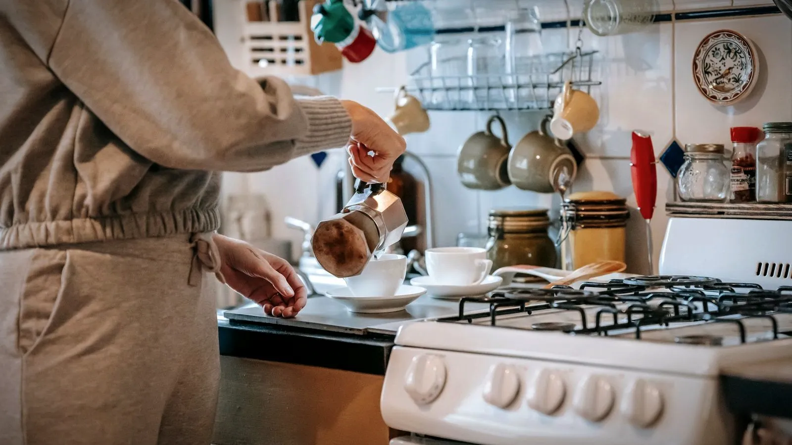 Person pouring freshly brewed coffee from a moka pot into a ceramic cup next to a kitchen stove